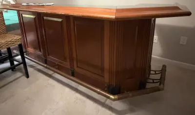 Large wooden home bar with a polished countertop, paneled front, and a brass footrest, showing some wear along the top edge.