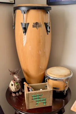 Latin Percussion Caliente conga drum and a smaller CP bongo drum, both featuring natural wood finishes, displayed alongside rustic...