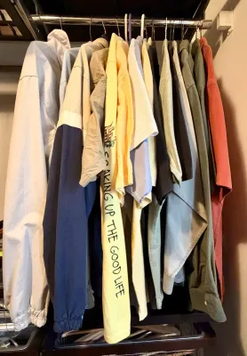 Assorted men's casual shirts and jackets hanging on a closet rod, including a yellow graphic long-sleeve shirt and a color-blocked...