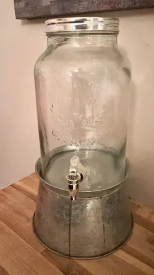 Large glass beverage dispenser with a silver-tone lid and spigot, resting on a galvanized metal stand.