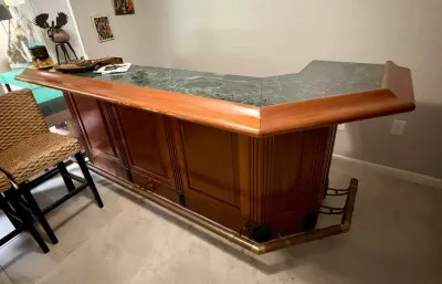 Mid-century style home bar featuring a green marble top, polished wood paneling, and a brass footrest, shown with a woven wicker barstool.