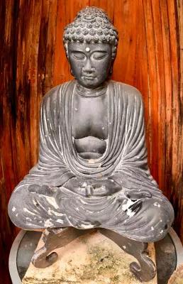 Seated Buddha statue in a dark grey finish, showing some weathering and wear on the lower portion, resting on a stone base.