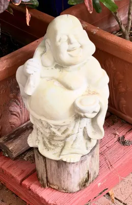 Weathered concrete laughing Buddha garden statue, showing natural outdoor patina and standing on a small wooden log pedestal.