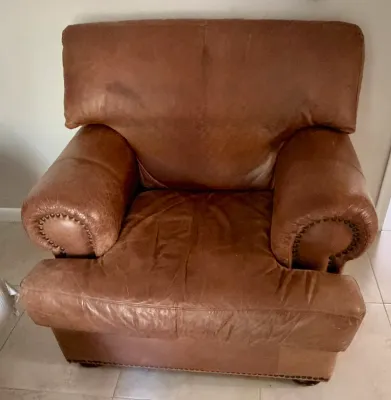 Overstuffed brown leather armchair featuring rolled arms with brass nailhead trim and a distressed, worn patina.
