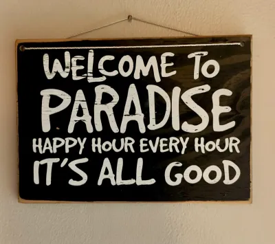 Rustic wooden hanging sign featuring white painted text on a black background reading "Welcome to Paradise, Happy Hour Every Hour,...