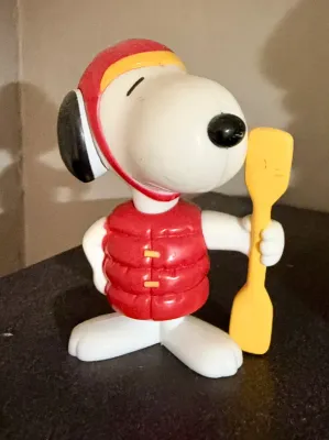 Vintage Snoopy plastic figure wearing a red life vest and helmet while holding a yellow paddle, showing minor surface wear.