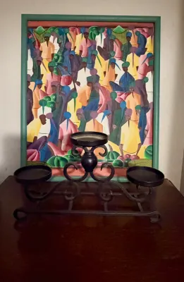 Haitian market scene painting in a green wooden frame, displayed behind a black wrought iron three-candle holder.