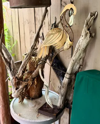 Stylized metal flamingo sculpture with a weathered finish, displayed alongside natural driftwood pieces on a round outdoor table.