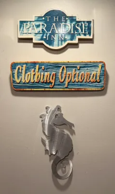 Trio of coastal-themed wall decor, including a blue and white "The Paradise Inn" sign, a colorful "Clothing Optional"...