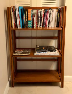 Mid-century modern style folding wooden bookshelf, featuring three shelves holding various books and magazines.