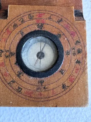 Sale Photo Thumbnail #56: An antique Chinese folding pocket sundial and compass (diptych style) crafted from wood. Features hand-painted Chinese characters, a central compass with a glass cover, and a folding metal gnomon on a hinged wooden plate. The underside and inner surfaces 