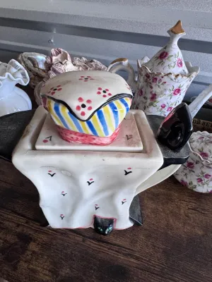 Sale Photo Thumbnail #177: A whimsical novelty ceramic teapot designed by Tony Carter, featuring a black cat with green eyes peeking out from under a draped white tablecloth. The lid is styled as a colorful striped and floral-patterned tea cozy or cake. Hand-painted details. Marked