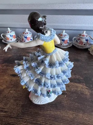 Sale Photo Thumbnail #96: A vintage porcelain figurine depicting a graceful ballerina in a yellow bodice and a tiered white and blue Dresden lace skirt. The delicate lace is accented with small applied porcelain roses. The base features elegant gold gilt scrollwork. Marked on the 