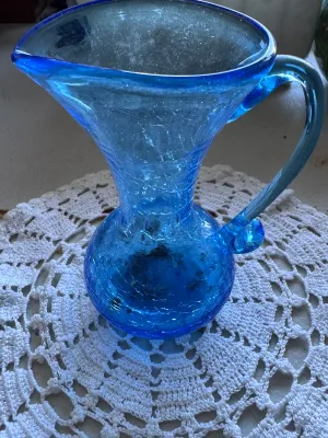 Sale Photo Thumbnail #331: A beautiful mid-century style hand-blown crackle glass pitcher or vase in a vibrant blue hue. Features a classic flared rim, a bulbous base, and an applied smooth glass handle. The piece shows characteristic crackle texture throughout the body. No maker's