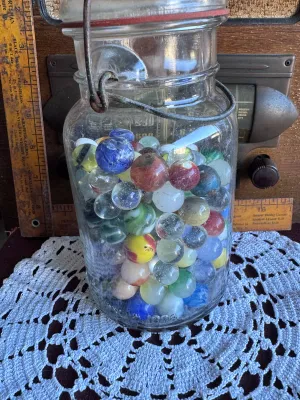 Sale Photo Thumbnail #81: A vintage clear glass canning jar with a wire bail closure and glass lid, filled with a large assortment of vintage glass marbles. The marbles feature various styles, colors, and sizes, including cat's eyes, solid colors, swirls, and clear shooters. The j