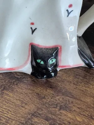 Sale Photo Thumbnail #180: A whimsical novelty ceramic teapot designed by Tony Carter, featuring a black cat with green eyes peeking out from under a draped white tablecloth. The lid is styled as a colorful striped and floral-patterned tea cozy or cake. Hand-painted details. Marked