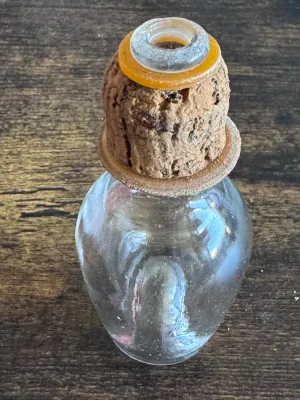 Sale Photo Thumbnail #234: A vintage miniature clear glass bottle, likely used for perfume, apothecary, or vanity display. Features a bulbous body shape with a cork stopper that includes a small glass ring top and amber-colored washer details. The glass shows some light age-related