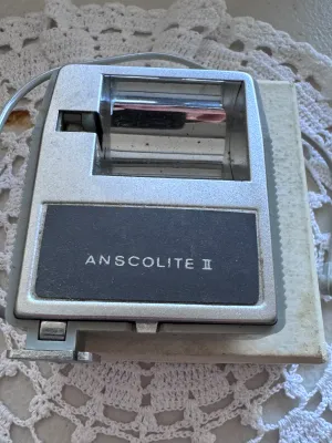 Sale Photo Thumbnail #315: Vintage Anscolite II camera flash unit manufactured by Ansco. Features a grey plastic housing with a built-in exposure guide dial on the back, a red test button, and an integrated PC sync cord. Marked 'Made in Japan' on the back casing. Includes the origi