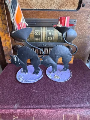 Sale Photo Thumbnail #303: A pair of vintage Halloween candle holders featuring arched-back black cats with yellow eyes on purple circular bases. Made of painted sheet metal or tin. One holder includes a partially burned orange, black, and white spiral candle, and the other contain