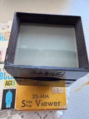 Sale Photo Thumbnail #313: Vintage photography lot featuring a Zarux 35mm Slide Strip Viewer (Royal De-Luxe model) made of dark bakelite/plastic, complete with its original cardboard box. The viewer is marked 'Zarux U.S.A.' on the bottom. Also included are two vintage boxes of Sylv