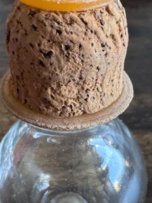 Sale Photo Thumbnail #236: A vintage miniature clear glass bottle, likely used for perfume, apothecary, or vanity display. Features a bulbous body shape with a cork stopper that includes a small glass ring top and amber-colored washer details. The glass shows some light age-related