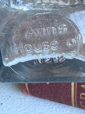 Sale Photo Thumbnail #256: A vintage clear glass jar featuring the embossed text "Ann's House of Nuts" on the bottom. The jar has a rectangular body tapering slightly towards the neck with a wide, rounded rim. It stands approximately 9.5 inches tall based on the adjacent ruler. Goo