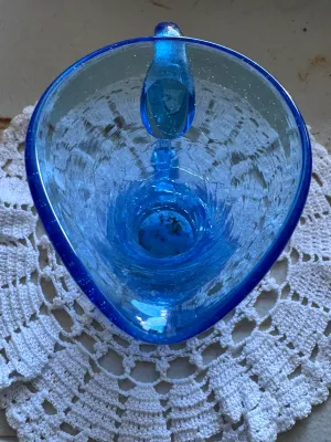 Sale Photo Thumbnail #334: A beautiful mid-century style hand-blown crackle glass pitcher or vase in a vibrant blue hue. Features a classic flared rim, a bulbous base, and an applied smooth glass handle. The piece shows characteristic crackle texture throughout the body. No maker's