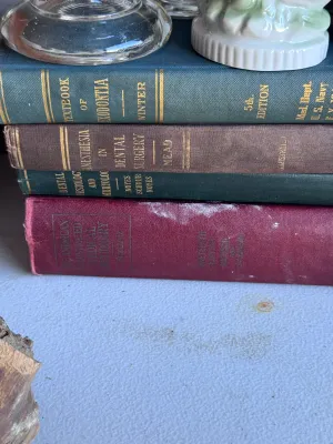 Sale Photo Thumbnail #172: A collection of 17 vintage medical and dental textbooks, primarily focusing on dentistry, oral surgery, and pathology. The hardback volumes feature classic cloth-bound covers in deep greens, blues, and burgundies with gilt lettering on the spines. Notable