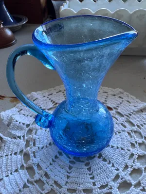 Sale Photo Thumbnail #332: A beautiful mid-century style hand-blown crackle glass pitcher or vase in a vibrant blue hue. Features a classic flared rim, a bulbous base, and an applied smooth glass handle. The piece shows characteristic crackle texture throughout the body. No maker's