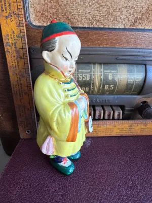 Sale Photo Thumbnail #196: A vintage hand-painted ceramic figurine depicting an Asian boy in traditional yellow and blue attire holding a banjo-like instrument. The base is stamped with a red maker's mark and reads 'Made in Occupied Japan', dating this piece to the post-WWII era (1