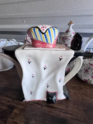 Sale Photo Thumbnail #176: A whimsical novelty ceramic teapot designed by Tony Carter, featuring a black cat with green eyes peeking out from under a draped white tablecloth. The lid is styled as a colorful striped and floral-patterned tea cozy or cake. Hand-painted details. Marked