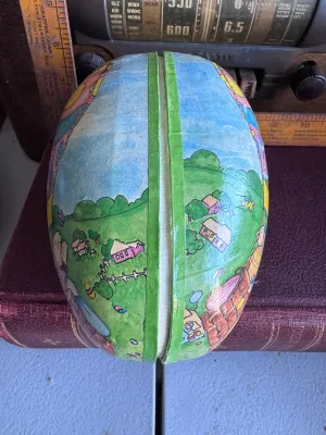 Sale Photo Thumbnail #191: A vintage large papier-mâché Easter egg candy container featuring a colorful illustration of bunnies in a hot air balloon on one side and a countryside landscape on the other. The interior is lined with a red floral pattern and features a white lace-sty