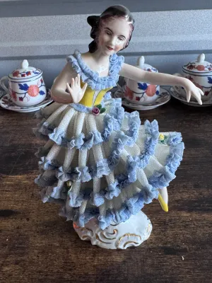 Sale Photo Thumbnail #91: A vintage porcelain figurine depicting a graceful ballerina in a yellow bodice and a tiered white and blue Dresden lace skirt. The delicate lace is accented with small applied porcelain roses. The base features elegant gold gilt scrollwork. Marked on the 