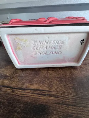 Sale Photo Thumbnail #231: A vintage novelty ceramic teapot shaped like a classic red AGA-style kitchen stove, featuring a sleeping tabby cat on top alongside a kettle and pot details. Marked on the underside 'Swineside Ceramics England'. Condition shows overall crazing to the glaz