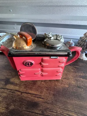 Sale Photo Thumbnail #230: A vintage novelty ceramic teapot shaped like a classic red AGA-style kitchen stove, featuring a sleeping tabby cat on top alongside a kettle and pot details. Marked on the underside 'Swineside Ceramics England'. Condition shows overall crazing to the glaz