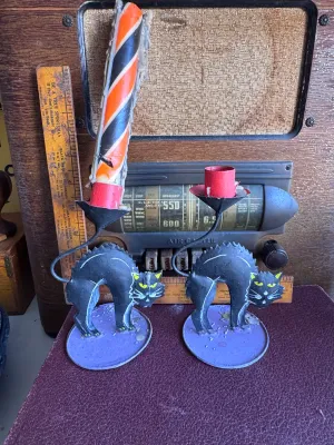 Sale Photo Thumbnail #301: A pair of vintage Halloween candle holders featuring arched-back black cats with yellow eyes on purple circular bases. Made of painted sheet metal or tin. One holder includes a partially burned orange, black, and white spiral candle, and the other contain