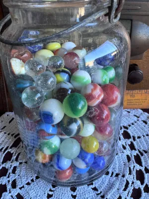Sale Photo Thumbnail #82: A vintage clear glass canning jar with a wire bail closure and glass lid, filled with a large assortment of vintage glass marbles. The marbles feature various styles, colors, and sizes, including cat's eyes, solid colors, swirls, and clear shooters. The j