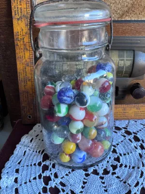 Sale Photo Thumbnail #80: A vintage clear glass canning jar with a wire bail closure and glass lid, filled with a large assortment of vintage glass marbles. The marbles feature various styles, colors, and sizes, including cat's eyes, solid colors, swirls, and clear shooters. The j