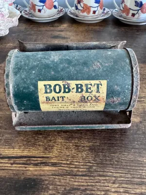 Sale Photo Thumbnail #222: A vintage Bob-Bet Bait Box featuring a cylindrical green metal body on a stand. The front label reads, "BOB-BET BAIT BOX - JUST HALF A TURN AND THERE'S YOUR WORM." The item shows significant vintage wear, including rust, paint loss, and surface grime cons