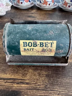 Sale Photo Thumbnail #223: A vintage Bob-Bet Bait Box featuring a cylindrical green metal body on a stand. The front label reads, "BOB-BET BAIT BOX - JUST HALF A TURN AND THERE'S YOUR WORM." The item shows significant vintage wear, including rust, paint loss, and surface grime cons