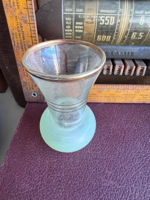 Sale Photo Thumbnail #299: A vintage small glass bud vase featuring a flared clear glass top section accented with gold-toned horizontal bands and a gold-gilded rim. The bulbous base is made of frosted light green glass, hand-painted with a delicate white flower and green leaves. M