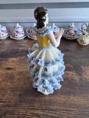 Sale Photo Thumbnail #95: A vintage porcelain figurine depicting a graceful ballerina in a yellow bodice and a tiered white and blue Dresden lace skirt. The delicate lace is accented with small applied porcelain roses. The base features elegant gold gilt scrollwork. Marked on the 