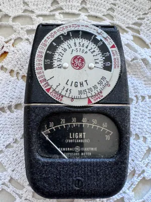 Sale Photo Thumbnail #308: A vintage General Electric Type DW-68 light exposure meter. This classic photographic tool features a black textured body with a dual-dial calculator on the front and a footcandle light scale. The back plate displays the serial number H66825 and is marked