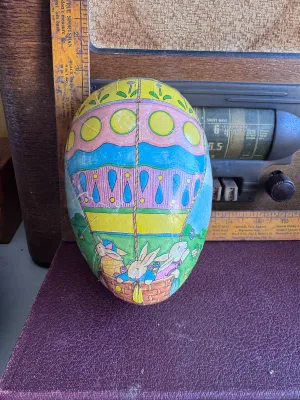 Sale Photo Thumbnail #192: A vintage large papier-mâché Easter egg candy container featuring a colorful illustration of bunnies in a hot air balloon on one side and a countryside landscape on the other. The interior is lined with a red floral pattern and features a white lace-sty
