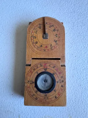 Sale Photo Thumbnail #59: An antique Chinese folding pocket sundial and compass (diptych style) crafted from wood. Features hand-painted Chinese characters, a central compass with a glass cover, and a folding metal gnomon on a hinged wooden plate. The underside and inner surfaces 