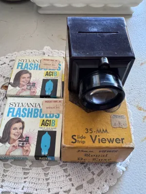 Sale Photo Thumbnail #310: Vintage photography lot featuring a Zarux 35mm Slide Strip Viewer (Royal De-Luxe model) made of dark bakelite/plastic, complete with its original cardboard box. The viewer is marked 'Zarux U.S.A.' on the bottom. Also included are two vintage boxes of Sylv