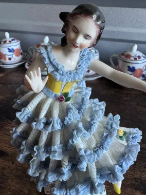 Sale Photo Thumbnail #92: A vintage porcelain figurine depicting a graceful ballerina in a yellow bodice and a tiered white and blue Dresden lace skirt. The delicate lace is accented with small applied porcelain roses. The base features elegant gold gilt scrollwork. Marked on the 