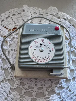 Sale Photo Thumbnail #314: Vintage Anscolite II camera flash unit manufactured by Ansco. Features a grey plastic housing with a built-in exposure guide dial on the back, a red test button, and an integrated PC sync cord. Marked 'Made in Japan' on the back casing. Includes the origi