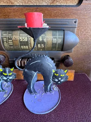 Sale Photo Thumbnail #302: A pair of vintage Halloween candle holders featuring arched-back black cats with yellow eyes on purple circular bases. Made of painted sheet metal or tin. One holder includes a partially burned orange, black, and white spiral candle, and the other contain