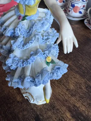 Sale Photo Thumbnail #93: A vintage porcelain figurine depicting a graceful ballerina in a yellow bodice and a tiered white and blue Dresden lace skirt. The delicate lace is accented with small applied porcelain roses. The base features elegant gold gilt scrollwork. Marked on the 